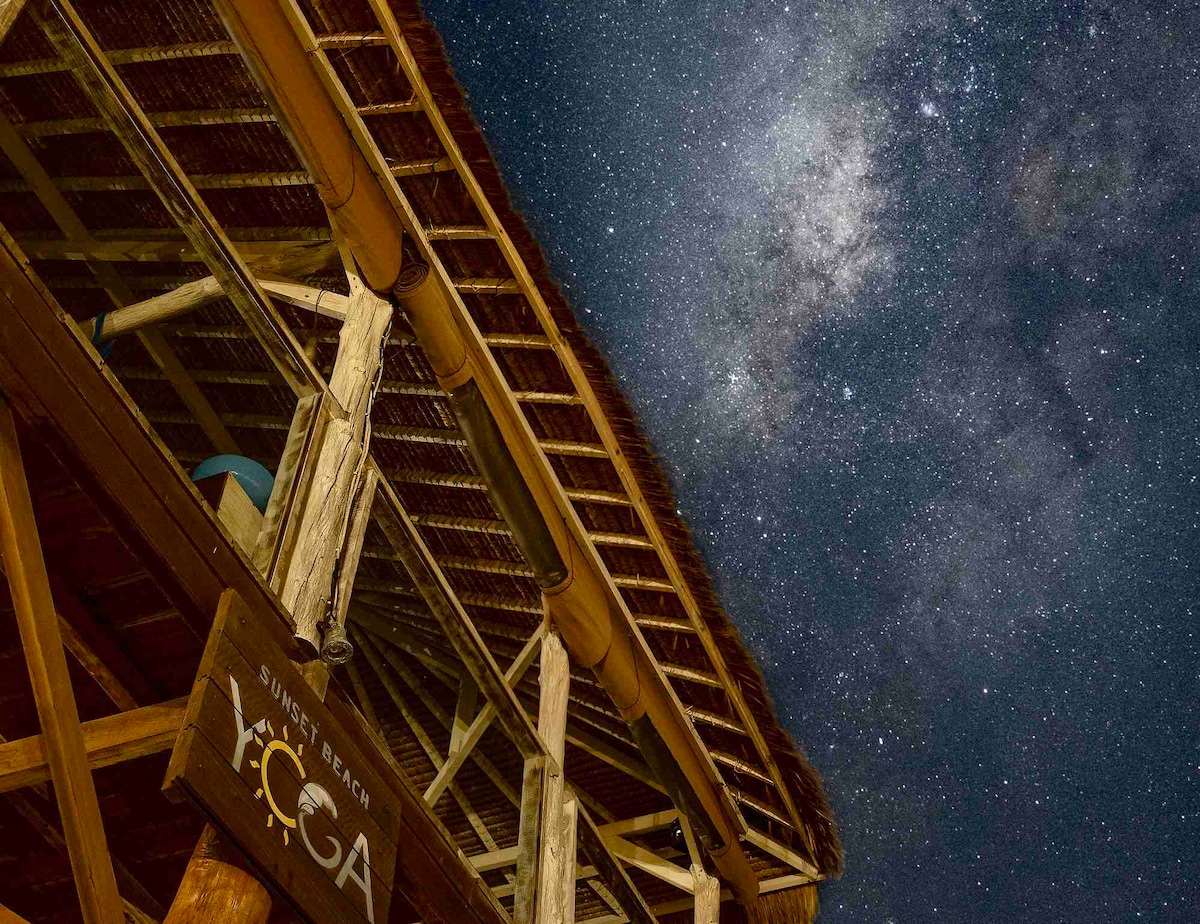 The structure of Sunset Beach Yoga is viewed from below, showcasing its natural materials and thatched roof under a starry night sky. The Milky Way is visible, adding to the serene atmosphere of the surrounding landscape.