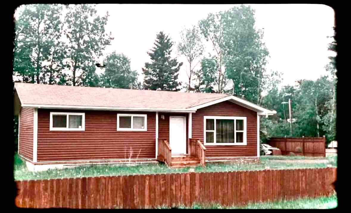 The exterior of a single-story home is shown, featuring earthy brown siding and a welcoming front porch with wooden steps. Large windows allow natural light to brighten the interior. A spacious lawn is surrounded by a wooden fence, and trees are visible in the background.