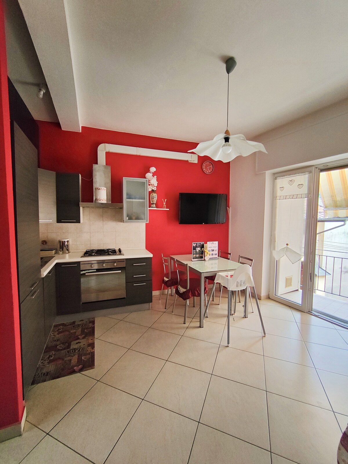 An open space kitchen and dining area features modern cabinetry against a red accent wall. A four-person dining table with metal chairs is positioned near a glass door, which provides access to a small balcony. A wall-mounted television completes the space.