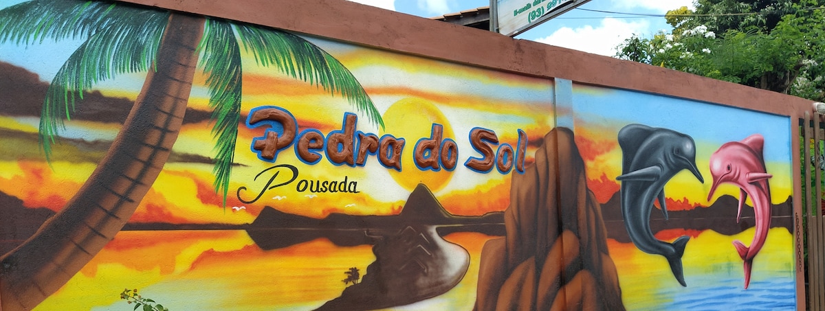 A vibrant mural depicts a sunset scene with an orange and yellow sky, mountains in the background, and stylized dolphins. The name 'Pedra do Sol Pousada' is prominently displayed in artistic lettering, creating an inviting focal point at the entry.