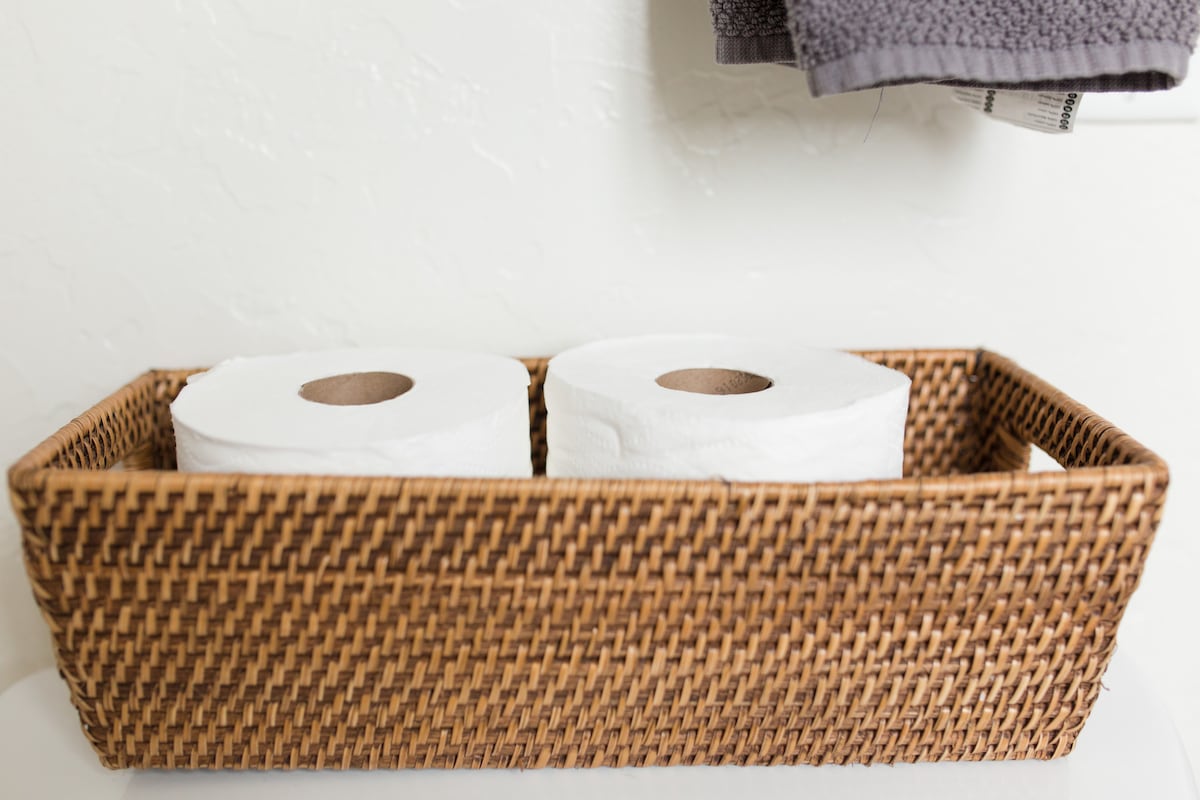 Two rolls of toilet paper are neatly arranged in a woven storage basket. The basket is placed on a flat surface, with a textured gray towel visible hanging above it. The wall features a light neutral color.