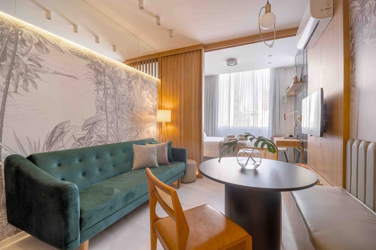 A modern living space features a teal velvet sofa, a round black table, and a light wood accent wall. Natural light filters through sheer curtains, highlighting a cozy atmosphere. A flat-screen TV is mounted on the wall, enhancing the functional design of the room.