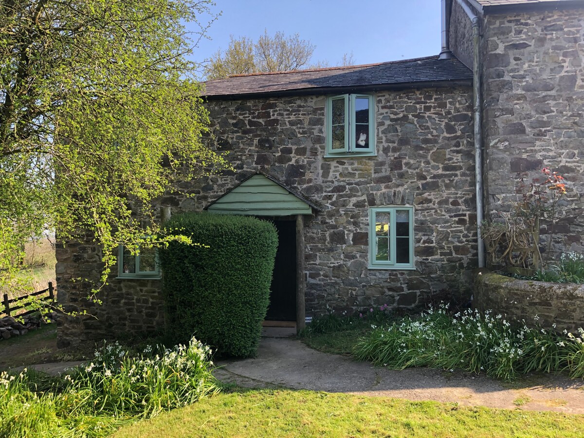 A charming stone cottage presents a rustic facade, framed by lush greenery and flowering plants. Two green windows and a welcoming door enhance the entrance, while a path leads through well-maintained grass. Surrounding fields and trees are visible in the background, contributing to the serene atmosphere.