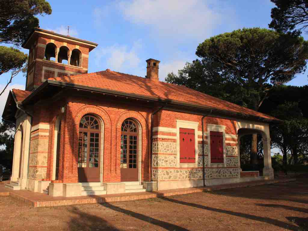 The exterior of the villa showcases a blend of brick and decorative stonework, featuring large arched windows and a small tower. The roof is covered with terracotta tiles, and the surrounding area includes tall pine trees, contributing to a serene and inviting atmosphere.