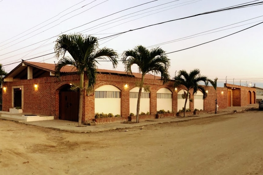 The exterior of the house features a warm brick facade, framed by palm trees. Large windows provide natural light, and a welcoming entrance is highlighted with a wooden door. The street is unpaved, contributing to a relaxed atmosphere.