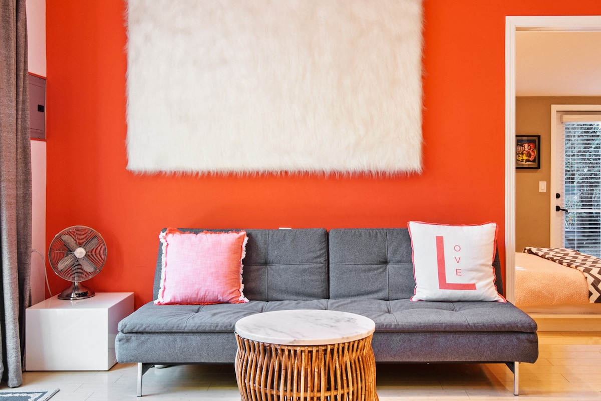 A sleek gray sofa is positioned against a vibrant orange wall, adorned with a large white textured artwork. Two decorative pillows accent the sofa, one featuring a red 'LOVE' design. A round table with a marble top and woven base sits in front of the sofa, with a small fan nearby.