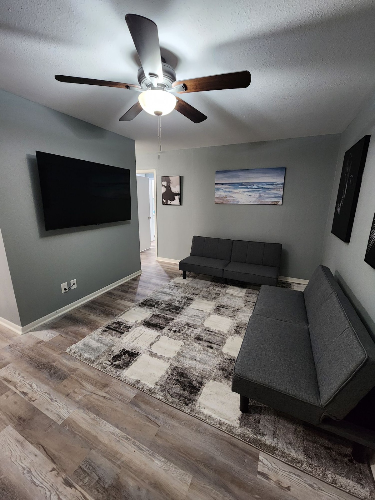 A modern living area is furnished with a gray sofa and a stylish area rug. A ceiling fan is visible above, providing airflow. Artwork adorns the walls, while a large flat-screen TV is mounted for entertainment. Natural light enters through the doorway.
