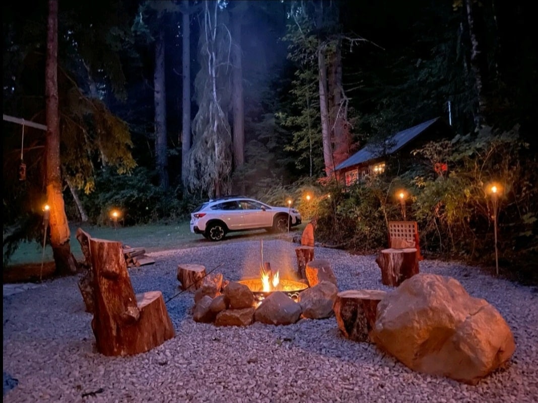 A secluded fire pit area is surrounded by handmade log chairs and large rocks, providing a cozy setting. Soft lighting from torches illuminates the space. In the background, a cabin discreetly blends with the forest, while a vehicle is parked nearby amidst the trees.