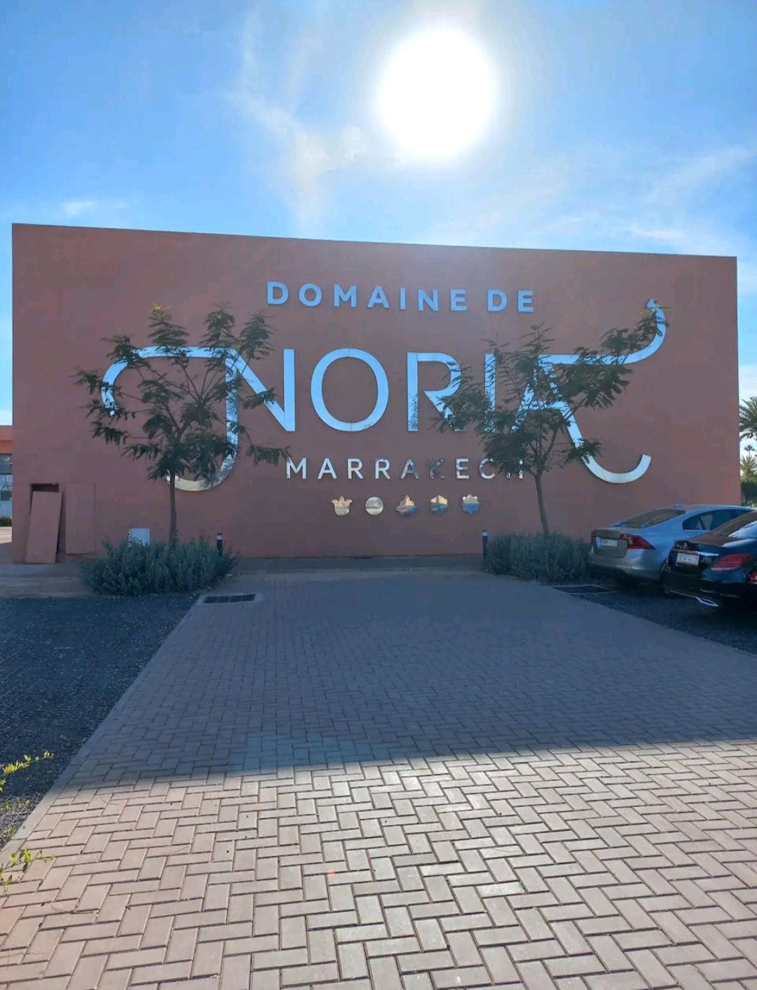 The entrance to Domaine de Noria is displayed on a large, peach-colored wall, showcasing the name in stylish lettering. Sunlight reflects off the wall, and the area is bordered by neatly arranged shrubs and a paved parking lot with several cars visible.