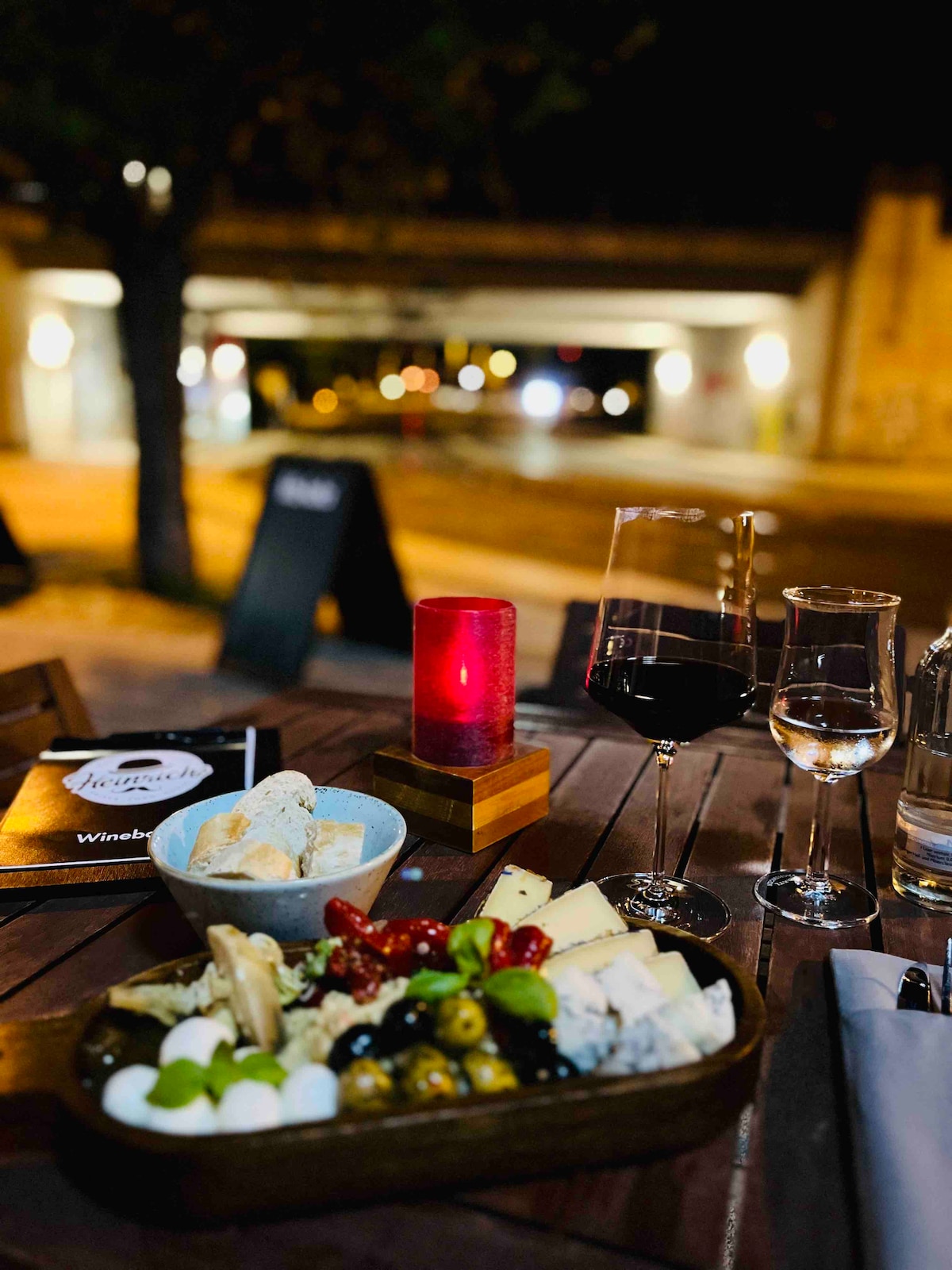 A table is set for an evening meal, featuring an assortment of cheeses and garnishes, with decorative glassware present. A lit candle adds a subtle glow, complementing two glasses of dark red wine and a menu nearby. Urban scenery is softly illuminated in the background.