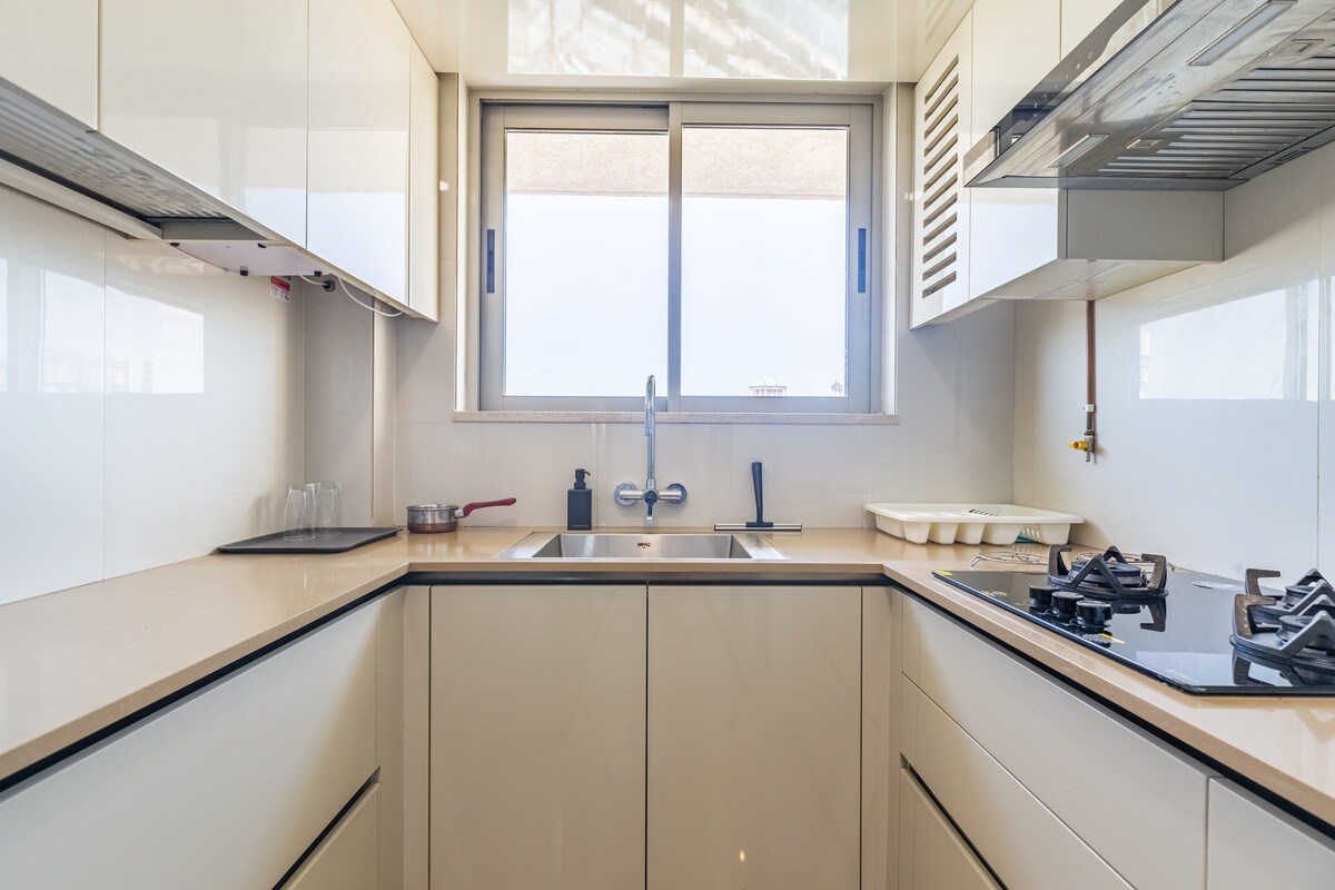 The kitchen is equipped with modern appliances, including a gas stove and washing area. Natural light enters through a window above the sink, illuminating the sleek countertops and cabinetry. Essential cooking items are neatly organized, providing a functional space for meal preparation.