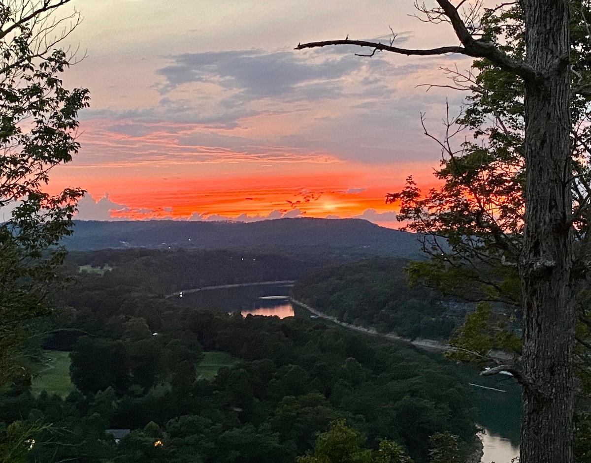 A breathtaking sunset is captured over rolling hills and a winding river. Vivid hues of orange and pink fill the sky, contrasting with the greenery of trees framing the scene. The tranquil landscape suggests a moment of serene beauty in the natural surroundings.