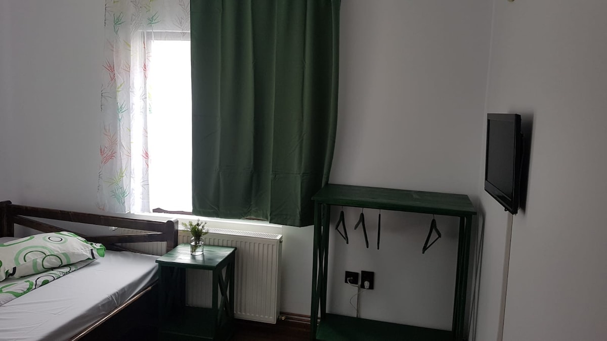 A bright room features a window dressed with a green curtain, allowing natural light to enter. A simple bed is accompanied by a nightstand on one side. A small TV is mounted on the wall above a green shelving unit, contributing to the room's functional design.