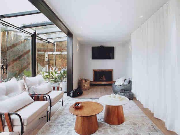 A light-filled living area features a comfortable sofa and two round wooden coffee tables. Floor-to-ceiling glass doors provide views of the outdoor space, while a wall-mounted TV is positioned above a cozy fireplace. Textured curtains add a soft touch to the room.