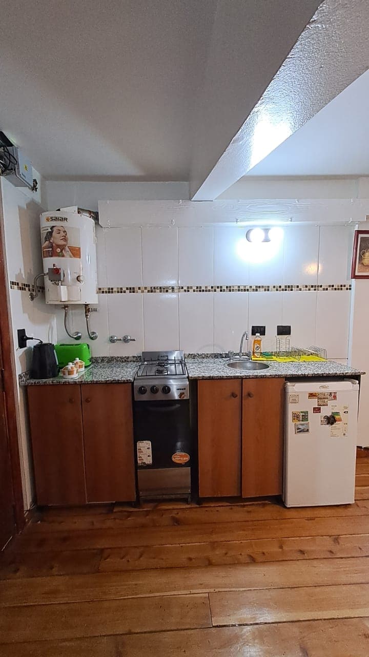 A functional kitchen area features granite countertops and wooden cabinetry. A stove, sink, and refrigerator are visible, along with small appliances and utensils on the counter. Warm wooden flooring adds to the inviting space.