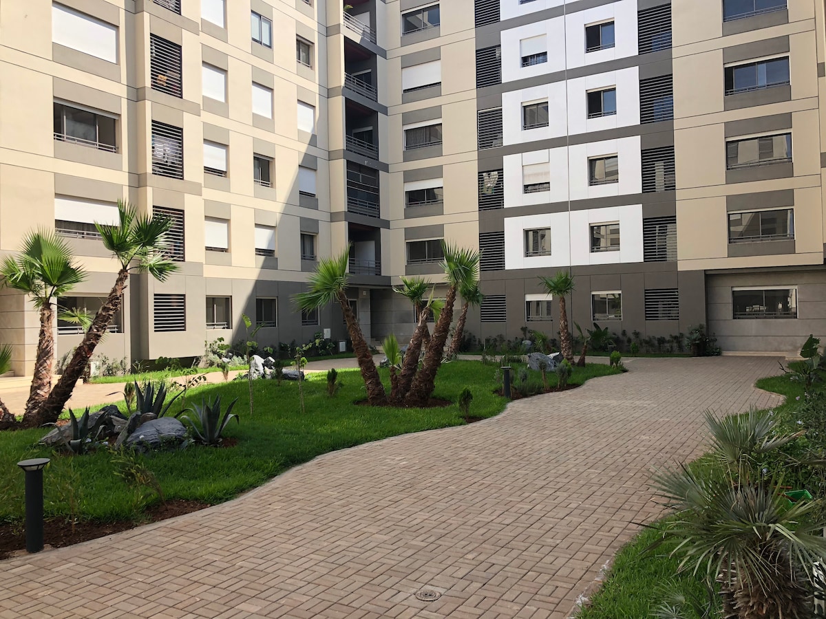A landscaped courtyard is framed by modern buildings, featuring a winding pathway lined with palm trees and manicured grass. Small rocks and shrubs add to the natural ambiance, while multiple windows reflect light throughout the space, enhancing the overall openness.