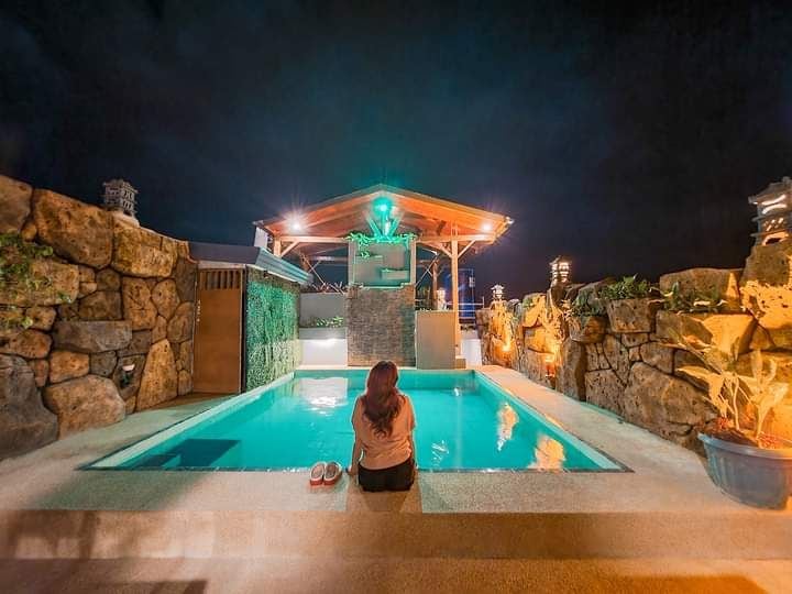 A tranquil pool area is illuminated at night, featuring a clear blue pool surrounded by natural stone walls and softly glowing lights. A covered structure is seen in the background, enhancing the relaxing ambiance. The scene evokes a serene atmosphere, ideal for unwinding.