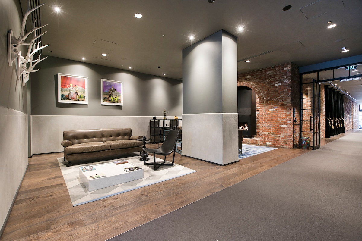 A modern lobby area is depicted with a cozy seating arrangement, featuring a spacious brown sofa and a stylish black chair on a patterned rug. Artwork adorns the walls alongside a brick feature wall, creating a warm and welcoming environment.