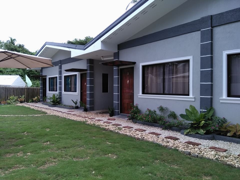 A modern exterior of the building is framed by well-maintained gardens, featuring colorful plants and smooth stone pathways. An umbrella provides shaded seating outside, while the structure's clean lines and large windows offer a welcoming appearance.