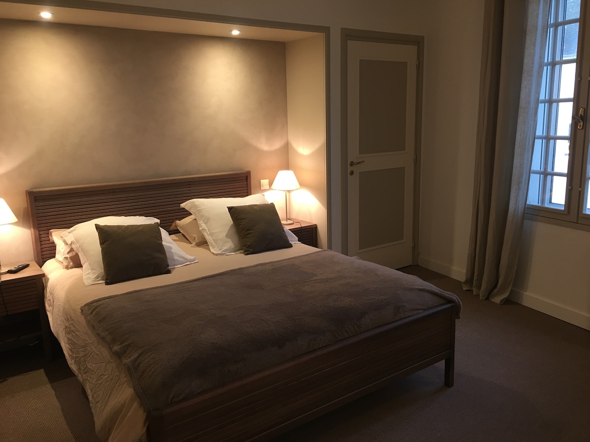 A spacious bedroom featuring a queen-size bed dressed in neutral-toned linens. Soft lighting is provided by bedside lamps, and a subtle color palette enhances the serene atmosphere. A window allows natural light to fill the room while providing a view of the outside.