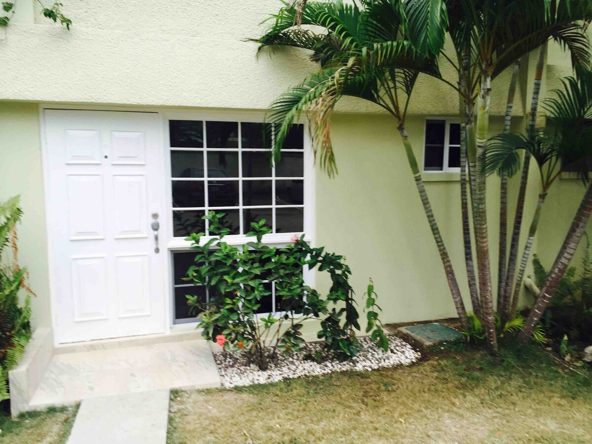 A welcoming entrance is framed by lush greenery and well-maintained landscaping. A white door with multiple glass panels stands at the forefront, accompanied by two adjacent windows. The light-colored exterior complements the vibrant plants, creating an inviting transition to the interior.