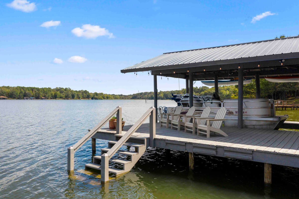 A serene lakeside dock is visible, featuring steps leading down into the water. Adirondack chairs are arranged on the dock, offering seating with a view of the calm lake. A covered area is present, providing shelter and space for watercraft. The surrounding landscape is lush and green.
