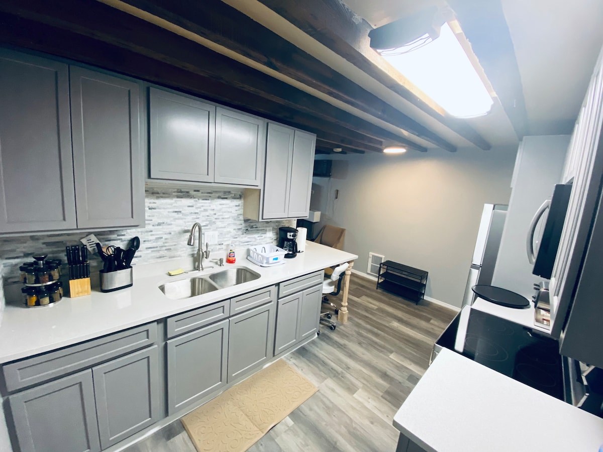 The kitchen features modern gray cabinetry and a spacious countertop. Stainless steel appliances, including a refrigerator and coffee maker, are visible. Textured tile backsplash complements the design. Wooden beams are present on the ceiling, adding character to the space.