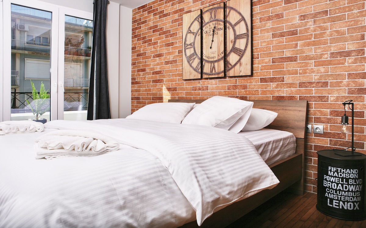 A spacious bedroom features a natural wood bed with a soft, light-colored striped duvet. A wooden clock adorns the brick wall, while a small green plant is placed on the bedside table. Soft light filters through the window, partially covered by dark curtains.
