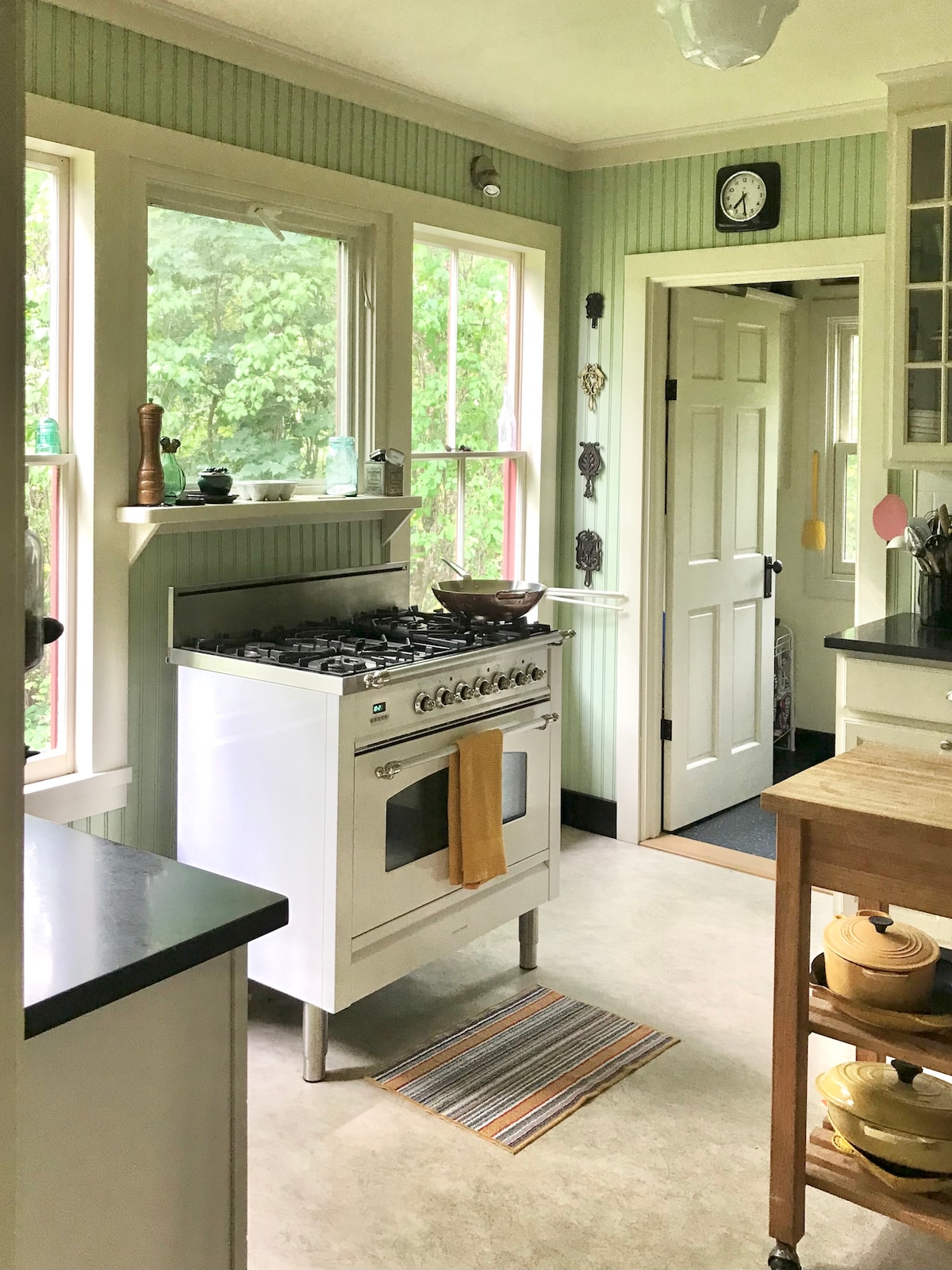 The kitchen features a white gas range with an oven, accented by colorful cooking utensils and a neatly organized countertop. Natural light flows through the multiple windows, illuminating the green bead-board walls. A door leads to a separate area, further enhancing the spacious layout.