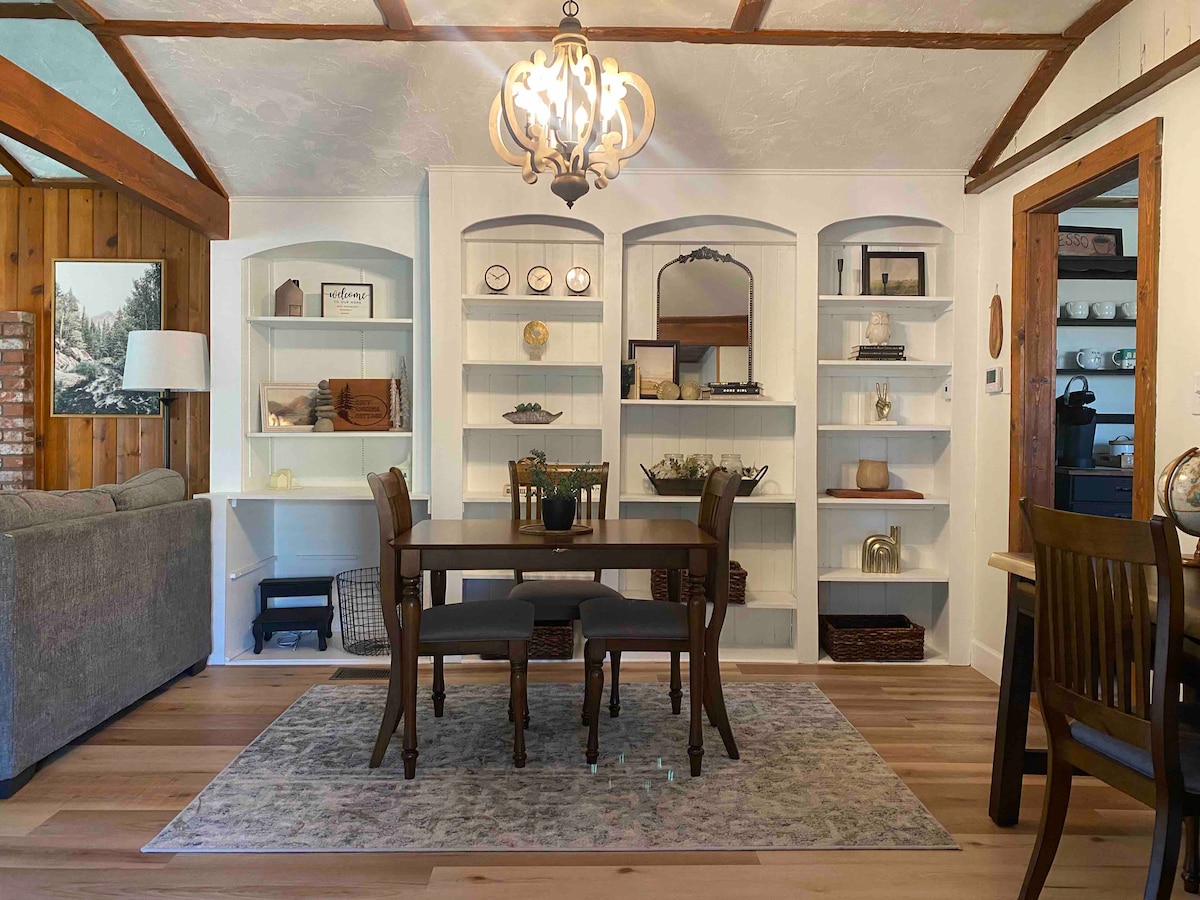 A dining area is featured with a wooden table surrounded by gray chairs. Shelving units display decorative items and organized storage, contributing to the space's functionality. Natural light enters through a nearby window, while a chandelier provides soft illumination from above.
