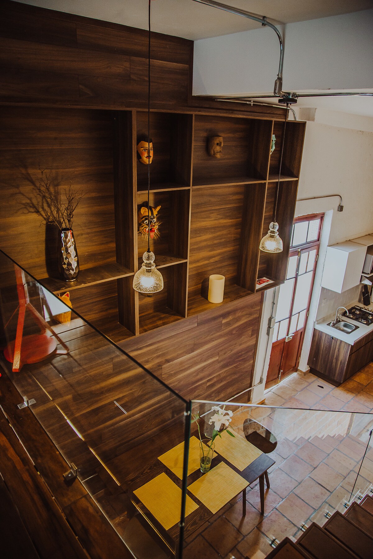 An open living space is highlighted by wooden accent walls and shelves displaying decorative items. Soft pendant lights illuminate the area, showcasing a small dining table with yellow placemats, next to a kitchen corner equipped with modern appliances.