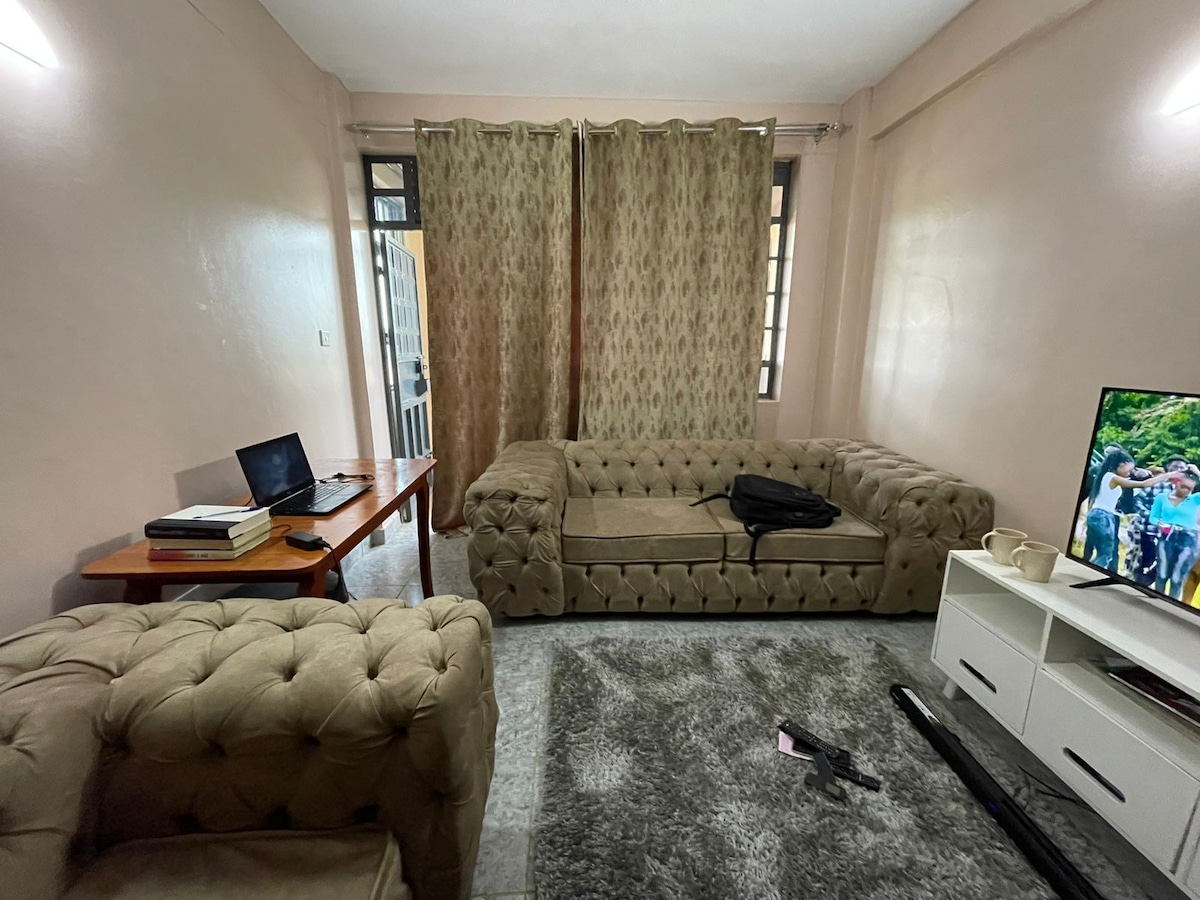 A comfortable living area features plush tufted sofas arranged for relaxation. Natural light filters through sheer curtains, illuminating the space. A wooden table with a laptop and books sits nearby, while a television displays a vibrant scene. A soft area rug adds warmth to the floor.