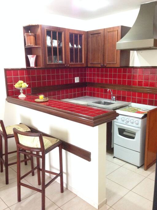 Cozinha americana com duas banquetas, frigobar, fogão quatro bocas, exaustor, pia de granito branco, armários de madeira.