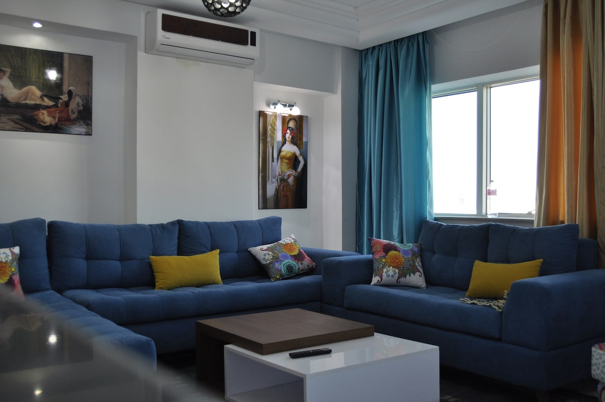A bright living room is featured, showcasing two blue sofas adorned with colorful pillows. A wooden coffee table sits centrally, with a remote control on its surface. Natural light enters through a window draped with light curtains, and a piece of artwork adorns one wall.