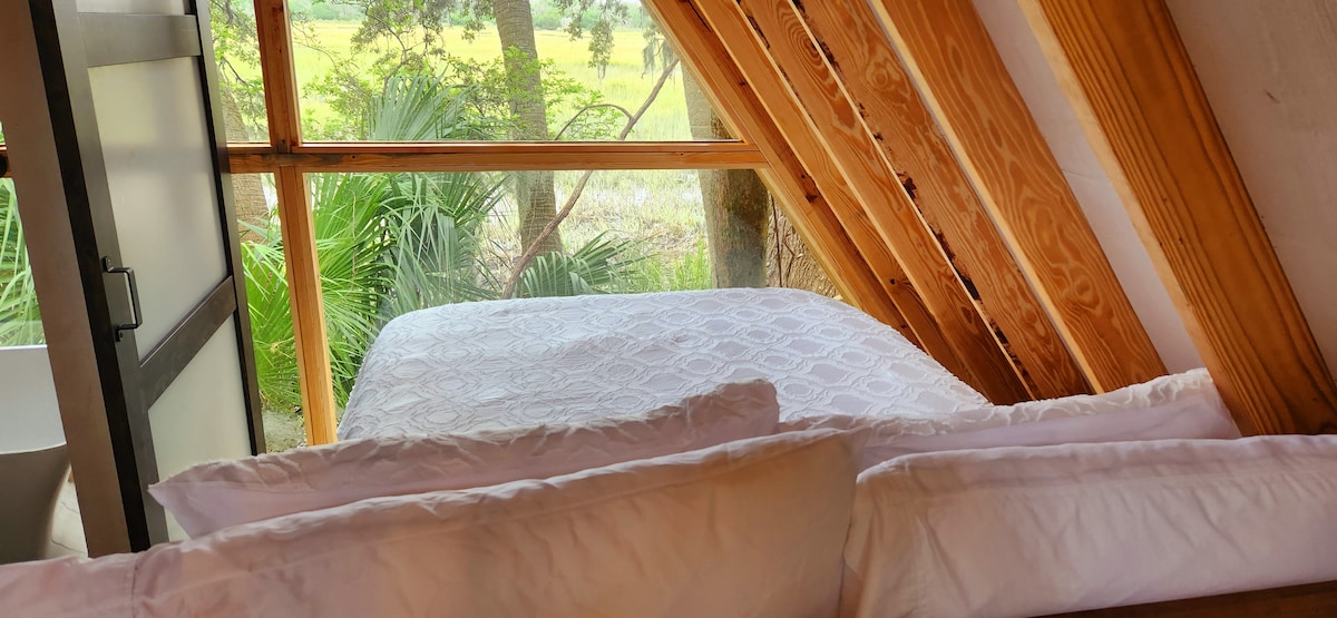 An open sleeping space features a spacious bed framed by natural wood beams. Large windows allow for ample natural light, providing views of the surrounding greenery and marsh. Soft white bedding and pillows create a serene setting for rest.