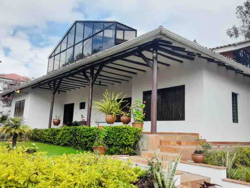 The exterior of a spacious home displays a modern design with large glass windows and a sloped roof. Lush green gardens surround the house, with various potted plants placed along the entryway. Steps lead up to a welcoming porch area, enhancing the inviting atmosphere.