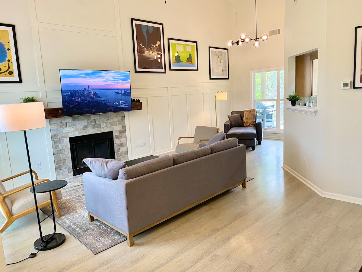 An open living area features a comfortable gray sofa facing a mounted television above a stone fireplace. Two accent chairs provide additional seating. Bright artwork adorns the walls, and natural light fills the space from large windows leading to a balcony.