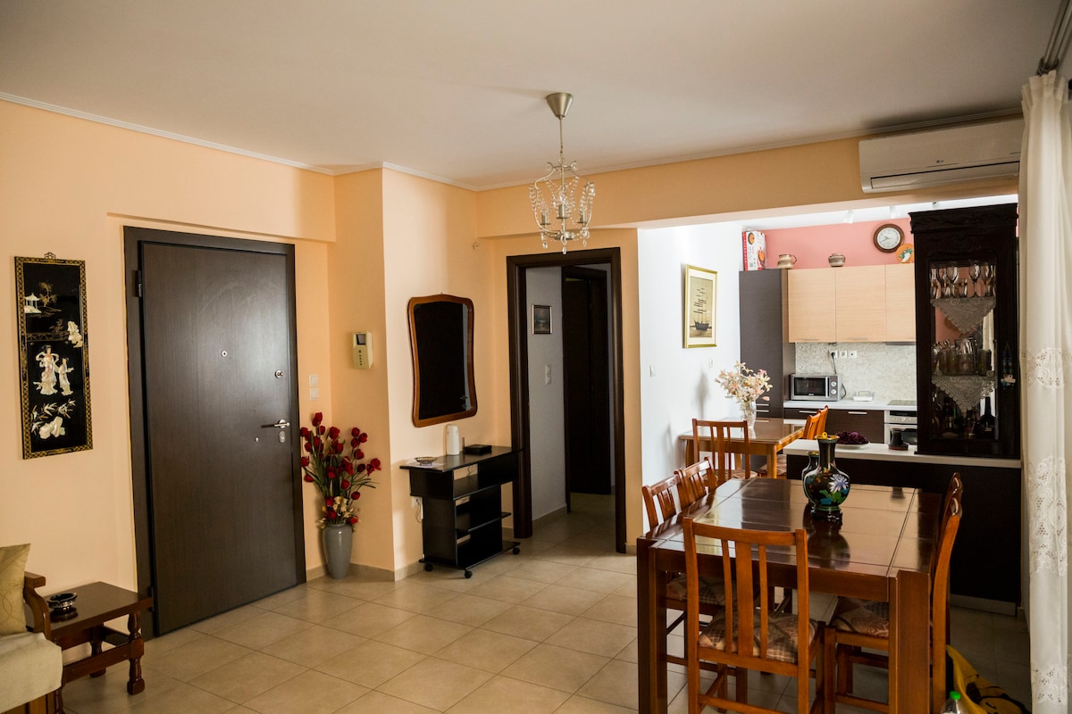 The living area features a dining table set for four, surrounded by wooden chairs. An adjacent kitchen is visible, equipped with modern appliances and cabinetry. A decorative mirror and a potted plant enhance the space, while warm wall colors contribute to a friendly atmosphere.