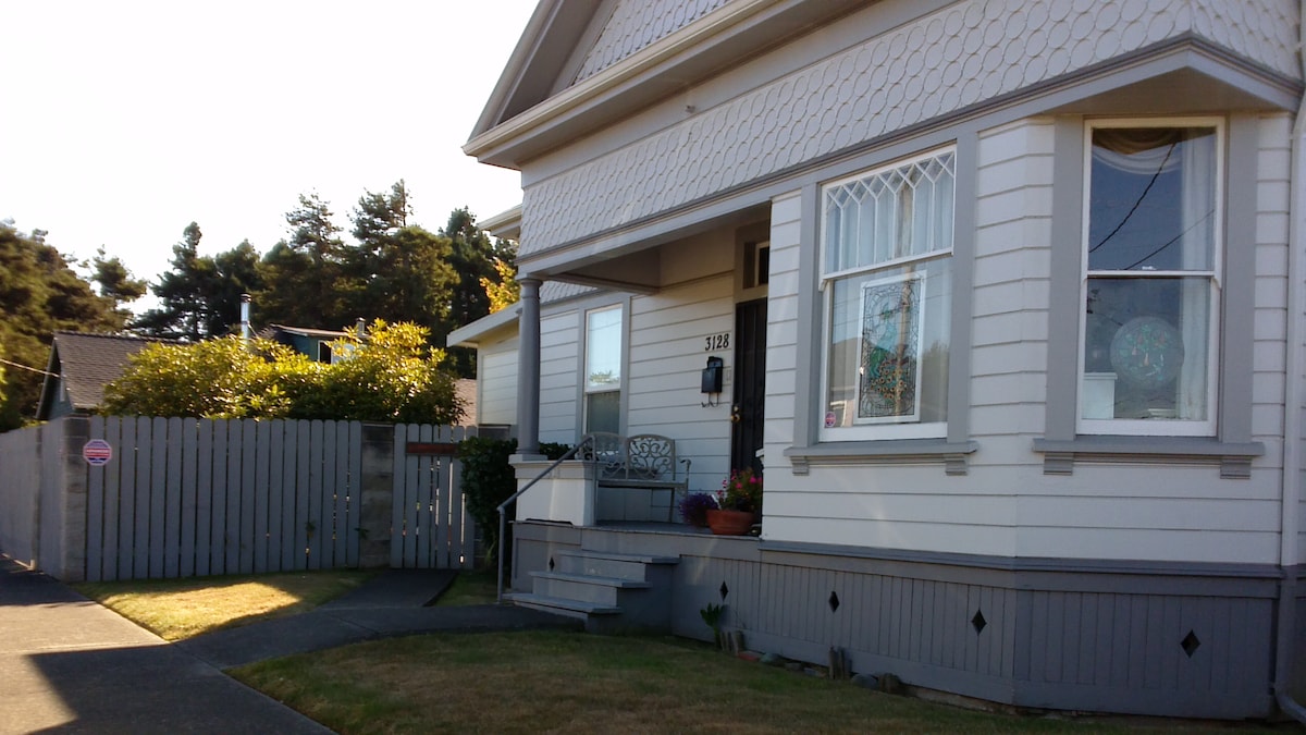 The exterior of a charming house is depicted, featuring a covered entryway with a small staircase leading to the front door. Large windows allow natural light to fill the space, and a wooden fence encloses the yard, which includes simple landscaping and a small seating area.
