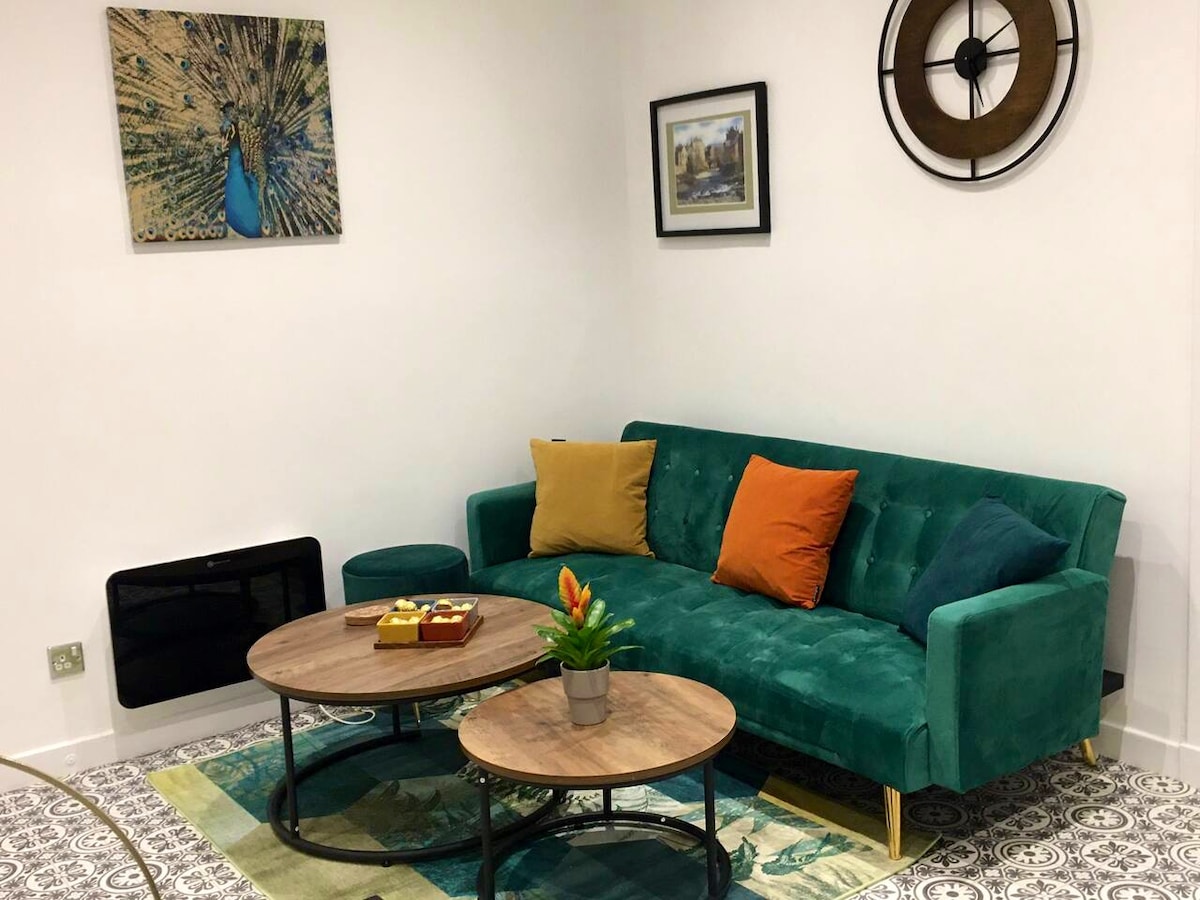 A stylish living area features a green sofa with colorful cushions and two round wooden coffee tables. A vibrant rug with intricate patterns is set on the floor, while a decorative wall clock and framed artwork enhance the space.