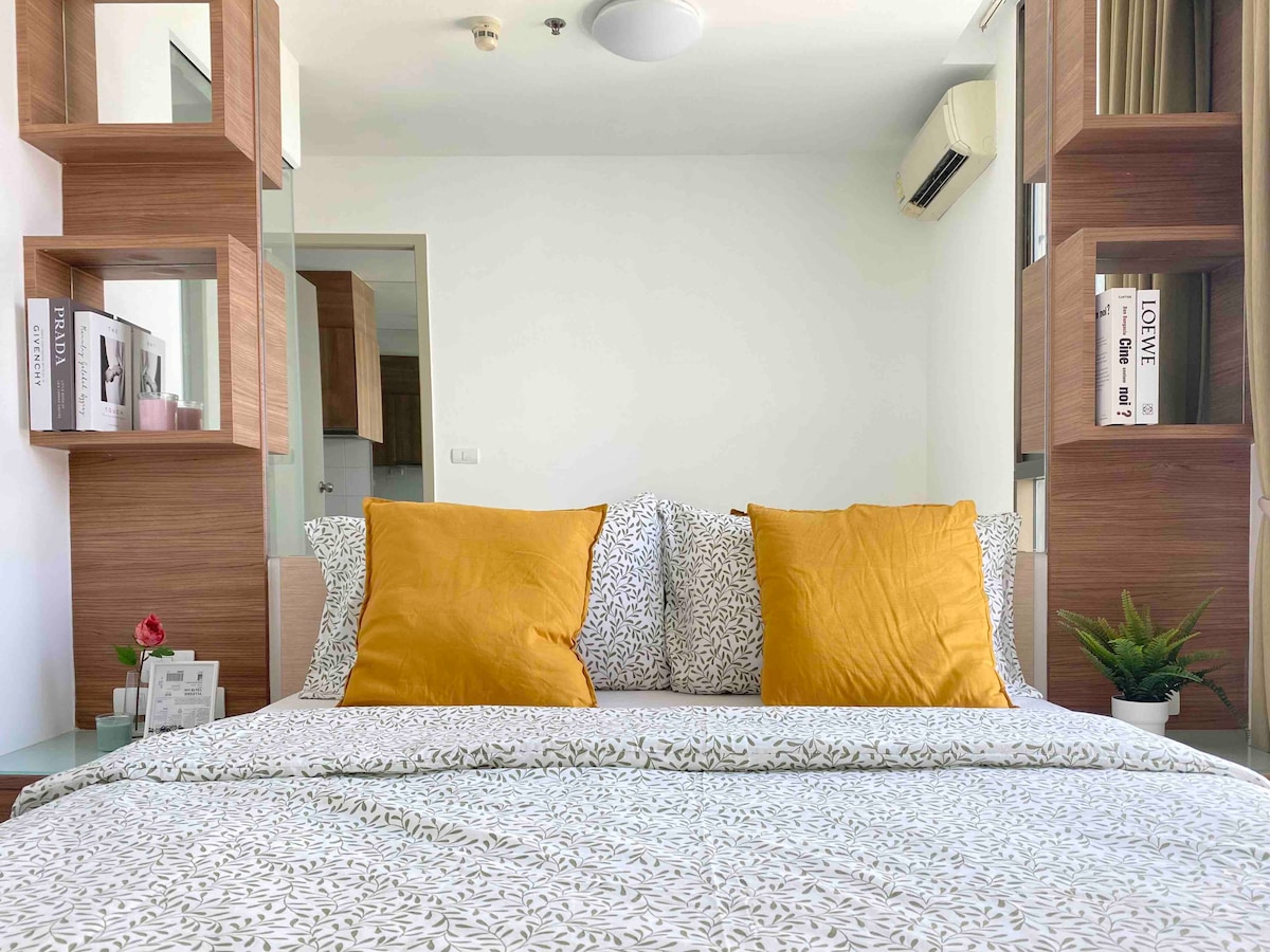 A spacious bed is dressed in a floral-patterned duvet and adorned with two large mustard-colored pillows. Surrounding shelves display books and decorative items, while natural light brightens the room through the window.