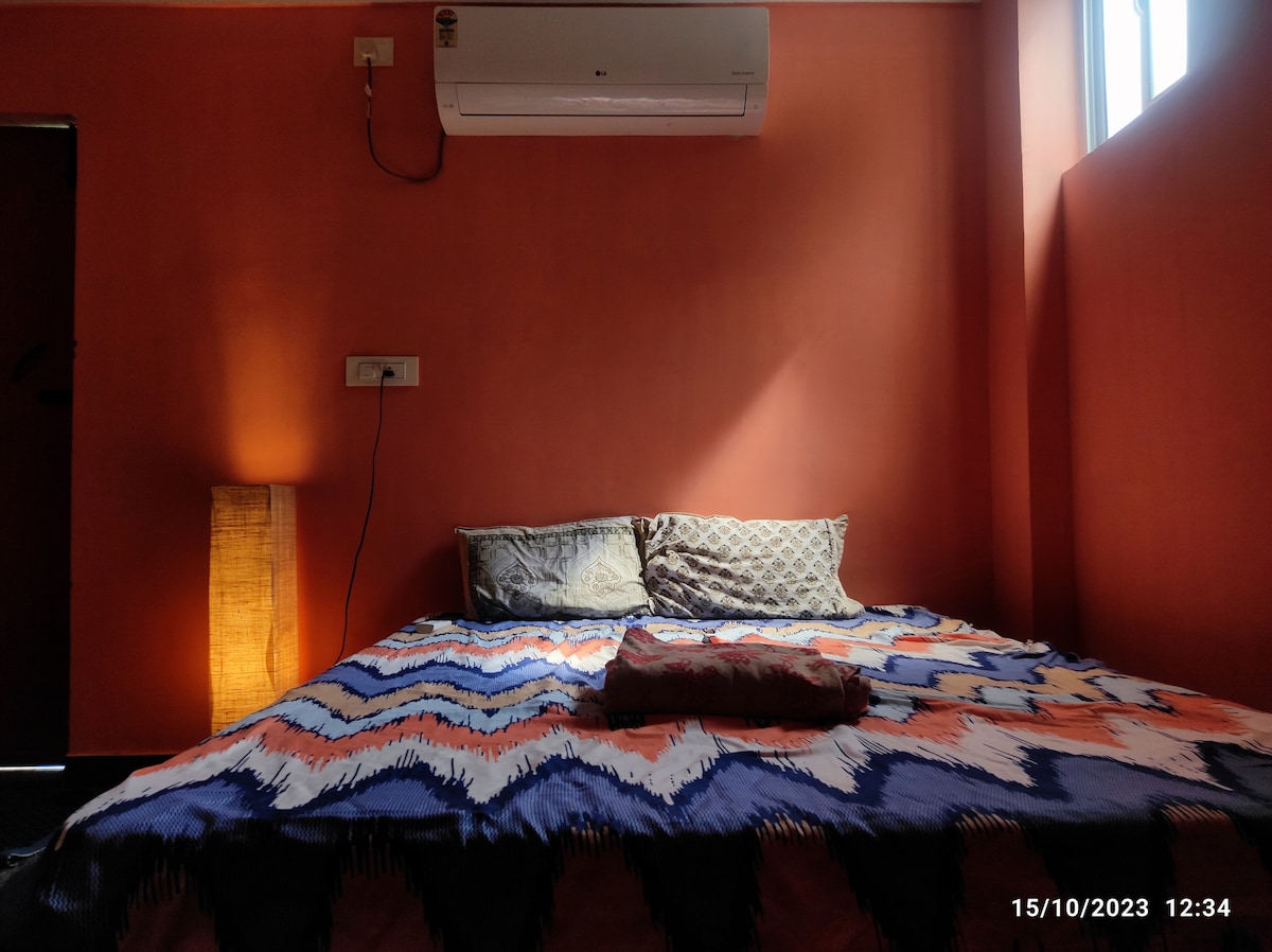 A comfortable sleeping area features a spacious bed adorned with a colorful patterned blanket. Alongside, a lamp provides soft light against a warm orange wall, complemented by two decorative pillows. A window allows natural light to highlight the inviting space.