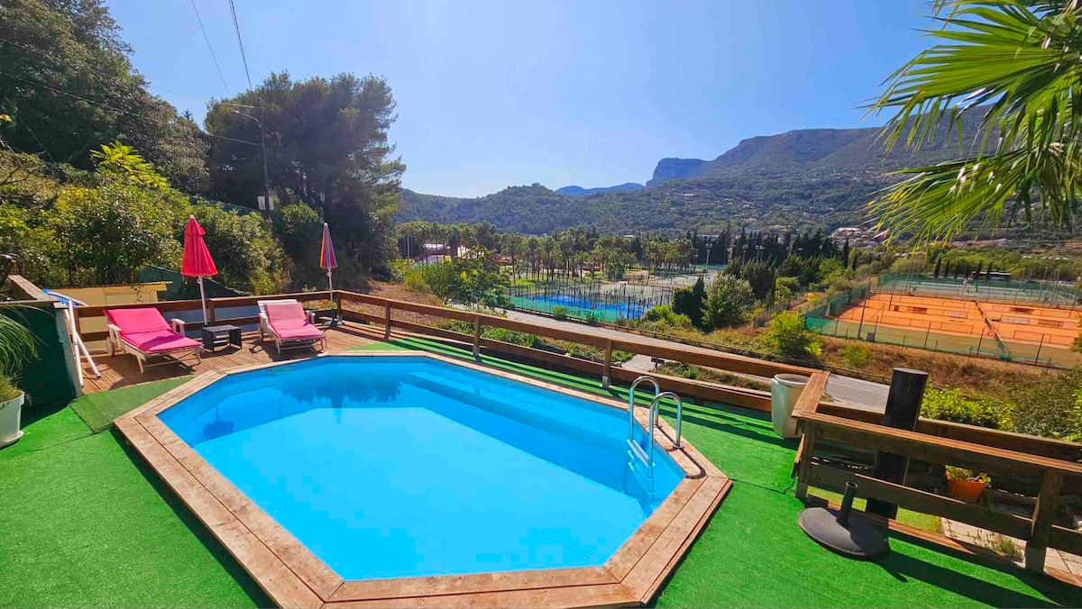 A private outdoor pool is surrounded by a wooden deck and greenery. Two pink lounge chairs are positioned beside the pool. In the background, tennis courts are visible, set against a scenic mountain view under clear skies.
