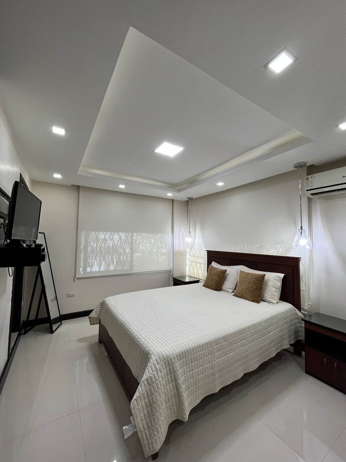 A spacious bedroom features a large bed with a textured white coverlet and two decorative pillows. Soft lighting is provided by recessed ceiling lights and pendant fixtures. A sleek television is mounted on the wall opposite the bed, and windows are fitted with light-filtering shades.