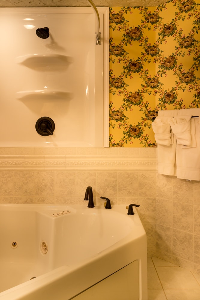A soaking tub is positioned adjacent to a shower enclosure, featuring floral wallpaper in soft yellow tones. A set of neatly arranged towels hangs on a rack nearby. The floor is tiled, complementing the serene atmosphere of the space.