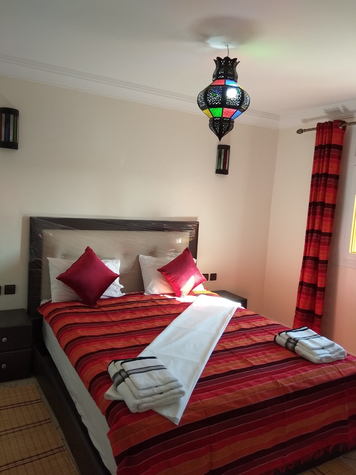 A cozy sleeping area features a spacious bed adorned with red and orange striped bedding. Decorative cushions are placed neatly on the pillows. Light is provided by a traditional lantern overhead, with colorful curtains framing the window, and towels are arranged thoughtfully on the bed.