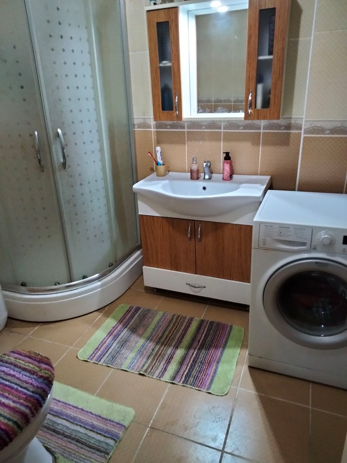 A shared bathroom is visible, featuring a glass shower stall, a compact vanity with wooden cabinetry, and a washing machine. Textured bath mats in vibrant stripes add color to the tiled floor, while essential toiletries are arranged on the vanity surface.