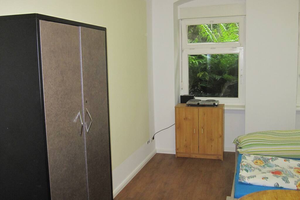 Schlafzimmer (Zi 2), Blick von Tür auf linke Seite des Zimmers mit Kommodenschrank am Fenster, drauf stehend 2 Kochplatten, dahinter 1 Toaster