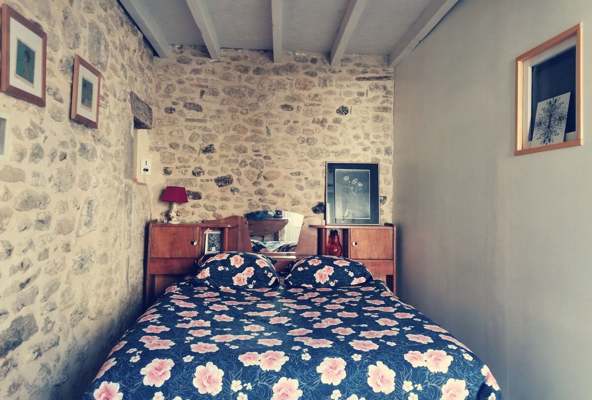 A cozy bedroom features a double bed adorned with a floral bedspread. Stone walls provide a rustic backdrop. A wooden nightstand is placed on either side of the bed, each featuring framed artwork and a lamp. Natural light enters through a small window.