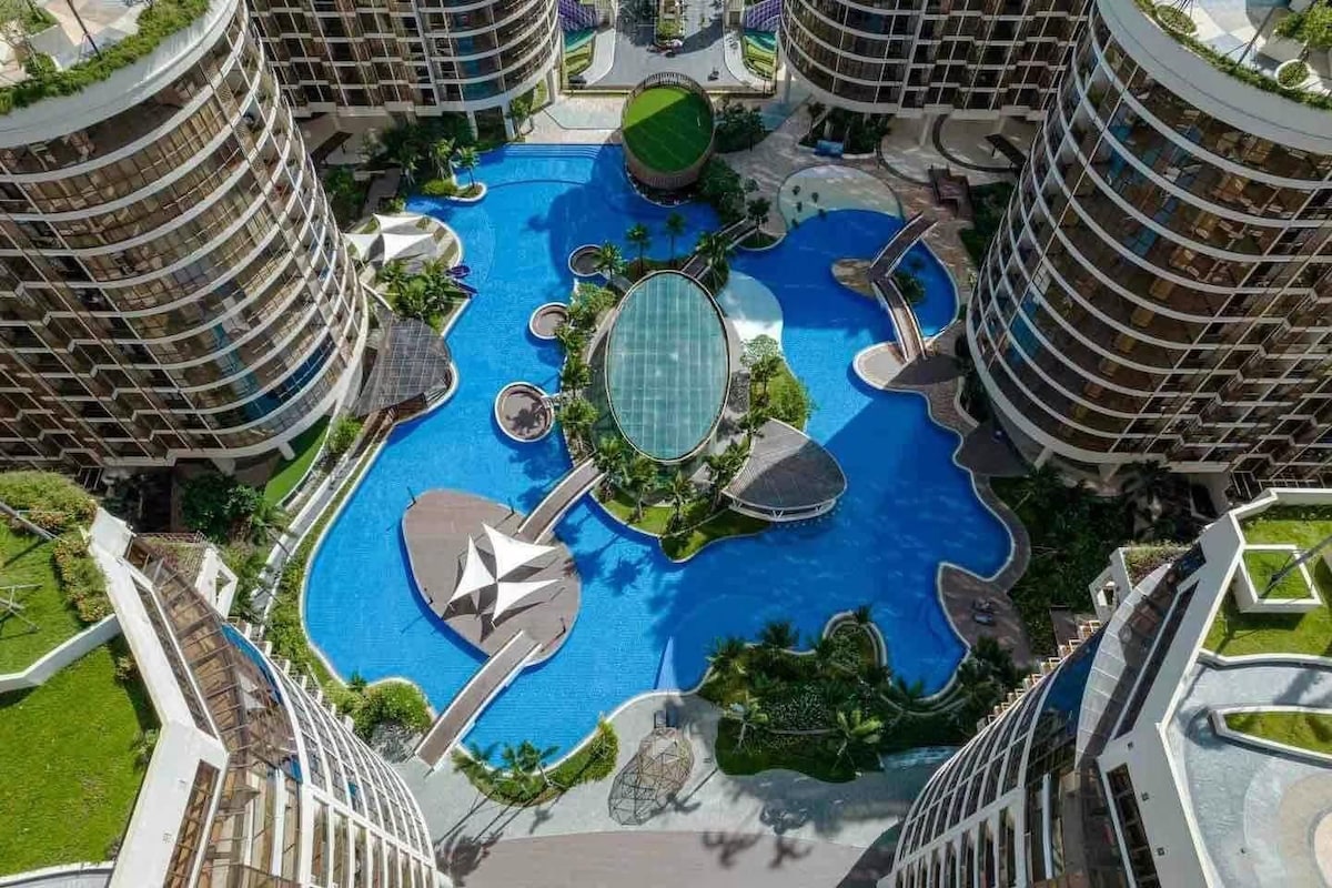 An aerial view captures a large, vibrant pool surrounded by lush landscaping, featuring curved shapes and various seating areas. Modern buildings with circular designs frame the pool area, creating a harmonious blend of architecture and nature.